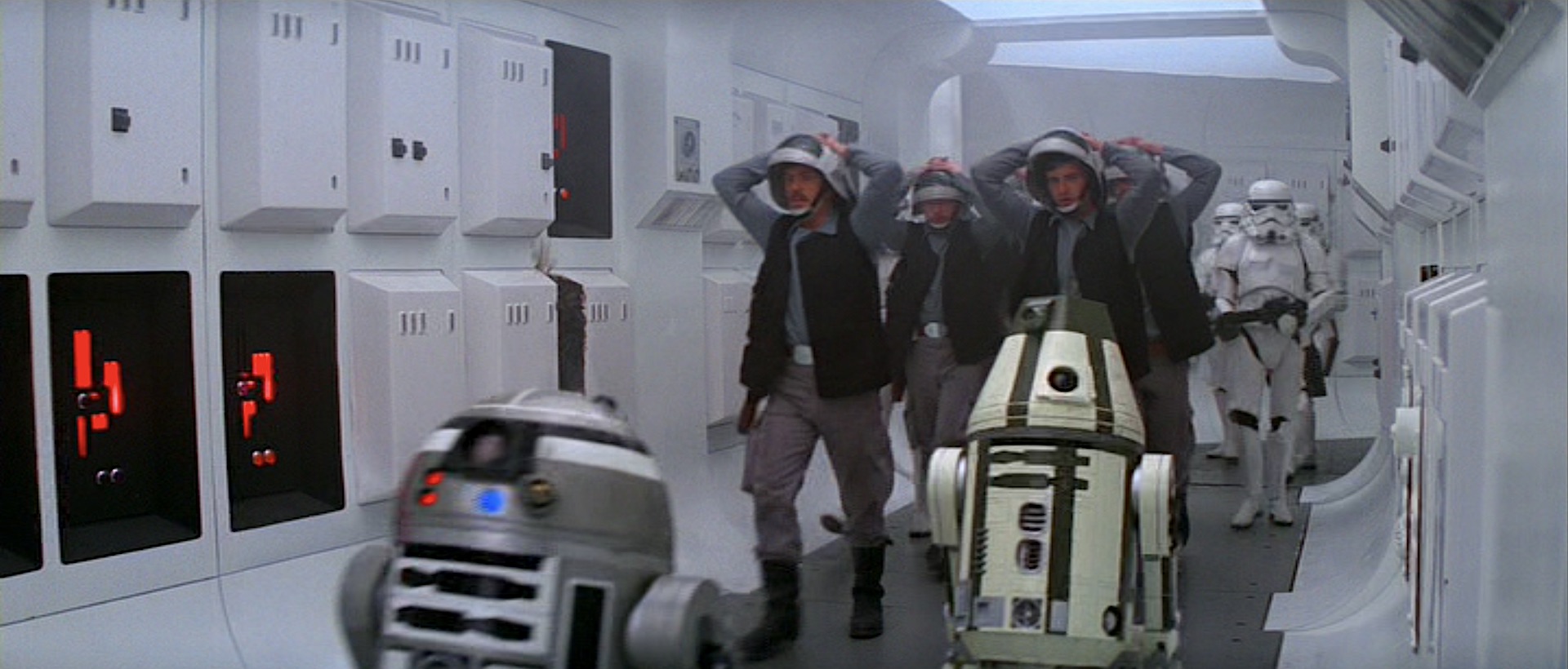 A still from Star Wars (1977) showing captured Rebel crew and several astromech droids being escorted by armed Imperial stormtroopers down a spaceship corridor.